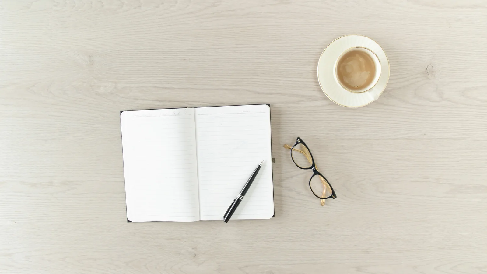 Vue de dessus d’une table de bois clair avec un carnet ouvert, un stylo, une tasse et des lunettes.